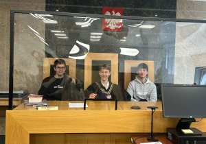 Troje uczniów pozują za stołem sędziowskim w sali rozpraw. Jeden z nich ma na sobie łańcuch sędziowski. Nad nimi widnieje godło Polski.