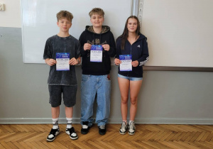 Troje uczniów stojących przed tablicą i prezentujących dyplomy konkursowe.