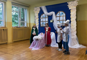 Młodzi aktorzy odgrywają scenę z bajki „Śpiąca Królewna” na tle dekoracji zamku.