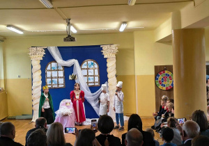 Dzieci w kostiumach odgrywają scenę z bajki „Śpiąca Królewna” przed zgromadzonymi rodzicami.