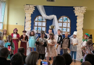Uczniowie stoją na scenie z tamburynami i innymi instrumentami perkusyjnymi podczas występu.