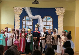 Grupa uczniów klasy 2b w kostiumach teatralnych stoi na scenie po zakończeniu przedstawienia.