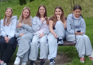 Pięć dziewcząt siedzi obok siebie na dużej drewnianej huśtawce zawieszonej na łańcuchach, umieszczonej na trawiastej skarpie. Cztery z nich mają na sobie luźne, szare dresy, a jedna z dziewcząt (druga z prawej) ubrana jest w ciemny T-shirt i spodnie dresowe. Dziewczęta uśmiechają się, rozmawiają i patrzą w stronę aparatu, jedna z nich trzyma telefon. Scena sprawia wrażenie relaksu i koleżeńskiej atmosfery na świeżym powietrzu.