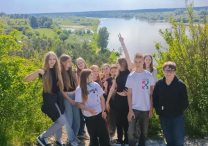 Grupa nastolatków pozuje do wspólnego zdjęcia na tle malowniczego krajobrazu z rzeką i zielonymi łąkami. Uczniowie stoją na skalistym wzniesieniu, wokół nich roślinność i krzewy. Część z nich uśmiecha się, jedna osoba unosi rękę w geście "victory", a pozostali patrzą w stronę aparatu. W tle rozciąga się panoramiczny widok na wodę, pola i lasy pod niebem z częściowo zachmurzonymi chmurami. Zdjęcie ma charakter pamiątkowy z wycieczki lub wyprawy szkolnej.