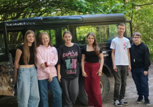 Grupa siedmiorga nastolatków stoi obok terenowego samochodu zaparkowanego na leśnej drodze. Pojazd znajduje się w cieniu drzew, a wokół widać gęstą zieleń. Młodzież jest ubrana na sportowo, w wygodne bluzy, koszulki i spodnie. Wszyscy pozują do zdjęcia, część z uśmiechem.
