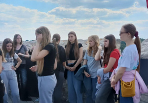 rupa nastolatek stoi na drewnianym tarasie widokowym, otoczonym kamiennym murem. Dziewczęta są ubrane w luźne, sportowe ubrania – głównie T-shirty i dresy – i rozmawiają ze sobą lub spoglądają w różne strony. W tle widoczne jest niebo z częściowo zachmurzonym błękitem. Po prawej stronie widoczny jest fragment czerwonej flagi.