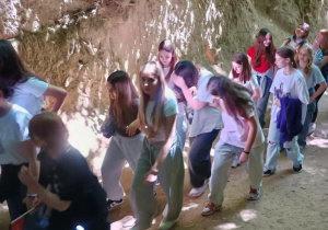 Grupa młodzieży słucha przewodnika w wąwozie lessowym, którego ściany porośnięte są korzeniami drzew wystających z górnej krawędzi urwiska. Uczniowie ubrani są w codzienne, wygodne ubrania – bluzy, koszulki i spodnie – i stoją w cieniu drzew, skupieni na opowieści prowadzącego. Przewodnik gestykuluje, a uczestnicy wycieczki stoją w półokręgu na tle jasnej, stromej ściany wąwozu. Atmosfera jest edukacyjna i zaciekawiona.