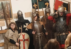 Zdjęcie wykonane wewnątrz historycznego pomieszczenia, gdzie kilku uczestników ubranych jest w średniowieczne stroje rycerskie i zbroje. Trzymają kopie i tarcze, a na ścianie za nimi widnieją herby, krzyż z wizerunkiem Chrystusa i średniowieczne insygnia. Z przodu kadru widać plecy siedzących uczestników, którzy obserwują scenę – prawdopodobnie w ramach lekcji muzealnej lub pokazowej inscenizacji historycznej. Atmosfera edukacyjna i tematyczna, związana z epoką średniowiecza.
