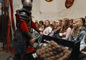 Zdjęcie przedstawia scenę edukacyjną wewnątrz muzeum historycznego. Rycerz w pełnej zbroi i czerwonym stroju prowadzi prezentację dla grupy młodzieży siedzącej w ławkach. Uczniowie uważnie słuchają i uśmiechają się. Na ławce przed nimi leży brązowy, pikowany kaftan ochronny oraz elementy uzbrojenia, w tym hełm. W tle widoczne są średniowieczne tarcze i herby zawieszone na ścianach.
