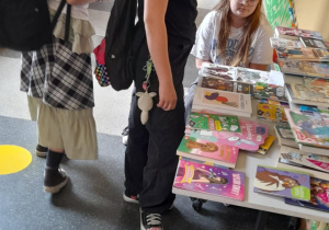 Kilka uczennic ogląda książki i czasopisma rozłożone na stole w szkolnym korytarzu. Jedna z dziewczynek siedzi za stołem, prawdopodobnie obsługując stoisko. W tle widoczne są kolorowe dekoracje, gazetki ścienne i plansze edukacyjne.