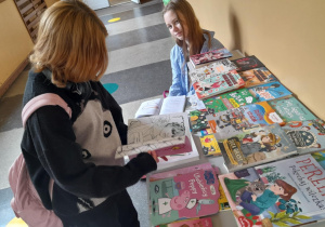 Dwie uczennice przy stole z książkami w szkolnym korytarzu. Jedna z dziewczynek siedzi i uśmiecha się, a druga przegląda książkę z czarno-białym rysunkiem. Na stole leży wiele kolorowych książek i publikacji dla dzieci i młodzieży.