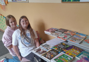 Dwie uśmiechnięte uczennice siedzą przy szkolnym stoliku, na którym ułożone są książki i czasopisma. W tle widać ozdobioną ścianę korytarza z gazetkami i dekoracjami z papieru.