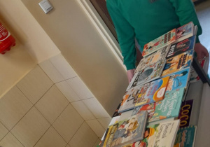 Uśmiechnięty chłopiec w zielonej bluzie stoi przy szkolnym stoliku z kolorowymi książkami. Na pierwszym planie widoczna jest ręka innej osoby sięgająca po książkę. W tle widać okno z doniczkami i część szkolnego korytarza.