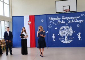 Dyrektor szkoły, pani Anna Michalak, wraz z uczniami Wiktorią i Emilem prowadzącymi uroczystość Miejskiego Rozpoczęcia Roku Szkolnego 2026/2027. W tle okolicznościowa dekoracja oraz biało-czerwone barwy z godłem Polski.