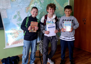 Trzech chłopców pozuje do zdjęcia na tle mapy Europy w klasie. Dwóch z nich trzyma w rękach kolorowe książki z serii 'Słownik', a jeden dyplom. W tle widać mapę, globusy na szafie oraz fragment szkolnej tablicy.