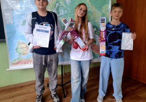 Trzech uczniów pozuje do zdjęcia w klasie na tle mapy Europy. Każdy trzyma nagrody: dyplomy, zestawy przyborów szkolnych i edukacyjne gry planszowe. W tle widoczna jest mapa ścienna oraz globusy na szafie.