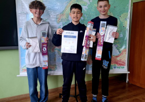 Trzech chłopców stoi na tle mapy Europy w klasie szkolnej. W rękach trzymają nagrody i dyplomy, prawdopodobnie za udział w konkursie geograficznym. Uśmiechają się i pozują do zdjęcia. W tle widoczna jest plansza z mapą oraz fragment tablicy i globus.