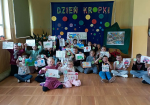 Grupa uczniów prezentuje swoje kolorowe prace przed dekoracją „Dzień Kropki” i Ogrodem Talentów.