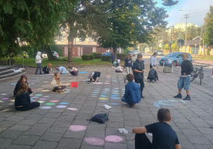 Uczniowie tworzą kolorowe rysunki kredą na chodniku przed szkołą podczas obchodów Dnia Kropki.