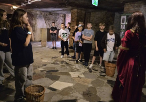 Grupa dzieci zwiedza podziemia z przewodniczką ubraną w historyczny strój. Dzieci stoją w szeregu na kamiennej posadzce w surowym, kamiennym wnętrzu z widoczną ceglaną ścianą i oświetleniem pod sufitem. Przed niektórymi stoją kosze wiklinowe.