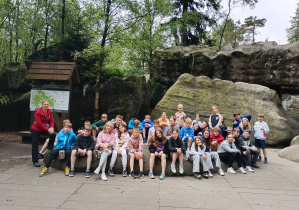 Grupa dzieci i dorosłych pozuje do zdjęcia na tle dużych, naturalnych skał w lesie. Większość dzieci siedzi na wielkim głazie, a z boku stoi kobieta w czerwonej kurtce. W tle widoczna jest tablica informacyjna Parku Narodowego Gór Stołowych oraz drzewa.