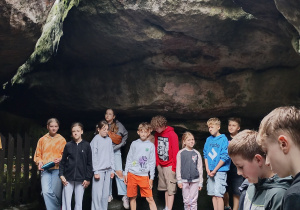 Grupa dzieci stoi pod dużym skalnym nawisem w parku przyrodniczym. Niektóre z nich patrzą przed siebie, inne trzymają telefony. Skały pokryte są mchem i wilgocią, a na ziemi widoczna jest kałuża z odbiciem.