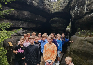 Grupa dzieci stoi na drewnianym pomoście wśród dużych formacji skalnych w otoczeniu lasu. W tle widoczne są drzewa, a niektóre dzieci opierają się o skały. Po prawej stronie na skale siedzi mały chłopiec. To zdjęcie z wycieczki przyrodniczej w terenie górzystym.