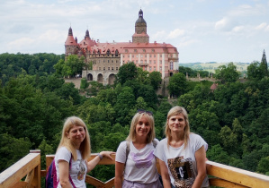 Trzy kobiety stoją na drewnianym tarasie widokowym, uśmiechając się do zdjęcia. W tle widoczny jest zamek Książ otoczony lasem, położony na wzgórzu. Nad zamkiem rozpościera się niebo z lekkim zachmurzeniem.