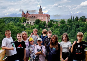 Grupa dzieci wraz z opiekunką pozuje do zdjęcia na tarasie widokowym. W tle widać zamek Książ otoczony zielonym lasem i wzgórzami. Dzieci stoją w rzędzie, niektóre się uśmiechają, jedna dziewczynka ma na głowie papierową koronę.