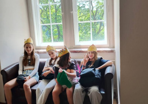 Cztery dziewczynki siedzą na brązowej sofie pod dużym, jasnym oknem. Mają na głowach papierowe korony i wyglądają na zadowolone. Za oknem widać zielone drzewa. Pomieszczenie ma drewnianą podłogę z geometrycznym wzorem.