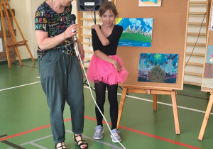 Nauczycielka trzymająca mikrofon rozmawia z dziewczynką ubraną w czarną bluzkę i różową tiulową spódniczkę. Stoją na sali gimnastycznej obok sztalugi z kolorowymi pracami plastycznymi. W tle widoczny jest głośnik i drabinki gimnastyczne.