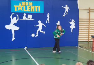 Dziewczynka w zielonej bluzie występuje solo na scenie podczas szkolnego wydarzenia ‘Mam Talent!’. Za nią znajduje się niebieskie tło z napisem ‘Mam Talent!’ oraz białymi sylwetkami przedstawiającymi różne pasje, m.in. taniec, grę na skrzypcach, piłkę nożną i jazdę na deskorolce. Na dole widać głowy siedzących widzów.