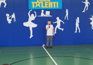 Chłopiec w białej koszulce i szarych spodniach stoi na scenie z mikrofonem w ręku podczas szkolnego wydarzenia ‘Mam Talent!’. Za nim znajduje się niebieskie tło z napisem ‘Mam Talent!’ i białymi sylwetkami przedstawiającymi różne pasje, takie jak taniec, malowanie, gra na skrzypcach i jazda na deskorolce. Na pierwszym planie siedzi publiczność złożona z dzieci.