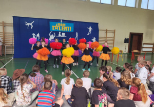 Grupa dziewczynek w kolorowych spódniczkach i z pomponami występuje na scenie podczas szkolnej imprezy ‘Mam Talent!’. Tło sceny jest niebieskie z żółtym napisem ‘Mam Talent!’ i białymi sylwetkami przedstawiającymi różne aktywności. Przed sceną siedzi liczna publiczność złożona z dzieci.