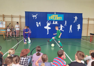 Trzech chłopców prezentuje umiejętności piłkarskie na scenie podczas szkolnego wydarzenia ‘Mam Talent!’. Dwóch ubranych w fioletowe stroje stoi przy piłce, trzeci w zielonym stroju wykonuje drybling. W tle niebieska dekoracja z napisem ‘Mam Talent!’ i białymi sylwetkami symbolizującymi różne pasje. Po lewej stronie siedzą trzy osoby przy sztalugach z rysunkami, a z przodu widoczna jest publiczność dziecięca.