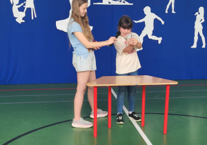 Dwie dziewczynki występują na scenie podczas szkolnego wydarzenia ‘Mam Talent!’. Jedna z nich trzyma mikrofon, a druga prezentuje wachlarz kart do gry, stojąc przy stole. W tle niebieska dekoracja z napisem ‘Mam Talent!’ i białymi sylwetkami symbolizującymi różne zainteresowania, takie jak taniec, malowanie czy gra na instrumencie.