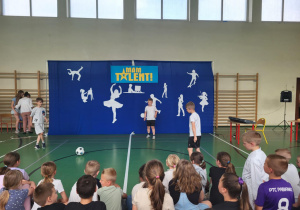 Chłopcy prezentują swoje umiejętności z piłką nożną na scenie szkolnego wydarzenia ‘Mam Talent!’. W tle znajduje się niebieskie tło z napisem ‘Mam Talent!’ i białymi sylwetkami symbolizującymi różne zainteresowania, takie jak taniec, gra na skrzypcach, malowanie czy deskorolka. Przed sceną siedzi publiczność złożona z uczniów.