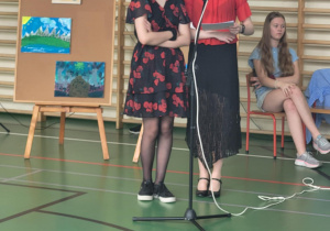 Dwie dziewczyny stoją przy mikrofonie na sali gimnastycznej, prowadząc szkolną uroczystość. Jedna z nich trzyma kartki z notatkami. W tle znajdują się sztalugi z kolorowymi pracami plastycznymi oraz uczniowie siedzący na krzesłach.
