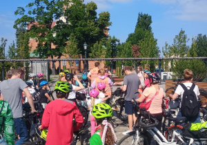 Grupa dzieci i dorosłych w kaskach rowerowych zgromadzona na placu z rowerami. W tle widoczny jest ceglany kościół z wieżą i otaczającymi go drzewami. Uczestnicy przygotowują się do wspólnego rajdu rowerowego w słoneczny dzień.