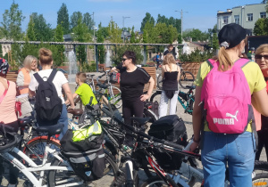 Grupa rowerzystów, w tym dzieci i dorośli, zgromadzona na placu z fontannami i ławkami. W tle widoczna jest zieleń, budynki mieszkalne i błękitne niebo z lekkim zachmurzeniem. Uczestnicy mają plecaki i rowery, przygotowując się do wycieczki.