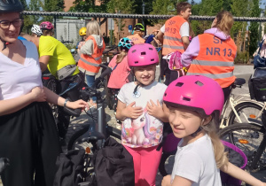 Dwie uśmiechnięte dziewczynki w różowych kaskach i różowych spodniach stoją przy rowerach podczas szkolnego rajdu rowerowego. W tle dorośli i dzieci w odblaskowych kamizelkach z napisem ‘SP NR 13’, a także kościół z wieżą i otaczające go drzewa.