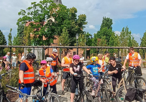 Grupa uczestników rajdu rowerowego zgromadzona na placu przed kościołem z wieżą. Dzieci i dorośli w kaskach i pomarańczowych kamizelkach odblaskowych przygotowują się do startu. Część osób trzyma rowery, wokół znajdują się drzewa, ławki i inni uczestnicy wydarzenia.