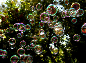 Na zdjęciu widoczne są liczne, unoszące się w powietrzu bańki mydlane, odbijające światło w różnych kolorach tęczy. Tło stanowi gęste, zielone liściaste drzewo oświetlone promieniami słońca,