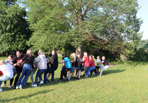 Na zdjęciu grupa dorosłych uczestniczy w zabawie w przeciąganie liny na trawiastym boisku. Uczestnicy, ustawieni w rzędzie, z zaangażowaniem ciągną linę w jednym kierunku, pochylając się do tyłu i trzymając ją obiema rękami. Na ich twarzach widać wysiłek i emocje. W tle widoczne są duże, zielone drzewa i słoneczna pogoda, która nadaje scenie radosny, piknikowy charakter.