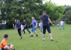 Na zdjęciu grupa dzieci i dorosłych gra w piłkę nożną na zielonym boisku. Uczestnicy są w ruchu – biegną, kopią piłkę i aktywnie uczestniczą w grze. Na pierwszym planie siedzi chłopiec w pomarańczowej koszulce, obserwujący mecz. W tle widać drzewa i ogrodzenie, a pogoda jest pochmurna, lecz jasna. Scena przedstawia wspólną, radosną aktywność ruchową na świeżym powietrzu.