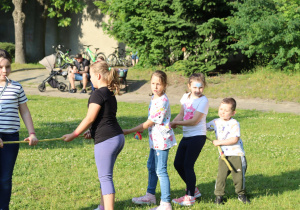 Grupa dzieci bawi się w przeciąganie liny na zielonej trawie w słoneczny dzień. Dzieci stoją w rzędzie, każde trzyma linę obiema rękami i ciągnie w jednym kierunku. W tle widać drzewa, ławkę, wózek dziecięcy i osoby siedzące, obserwujące zabawę. Jedna z dziewczynek ma na twarzy malunek, co świadczy o atmosferze festynu lub pikniku.