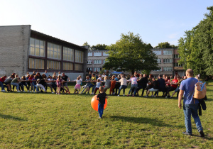 Na zdjęciu duża grupa dzieci i dorosłych bierze udział w przeciąganiu liny na zielonym boisku przy szkole. Uczestnicy stoją w dwóch rzędach po przeciwnych stronach liny, pochylając się i wkładając dużo siły w zabawę. W tle widać budynek sali gimnastycznej i szkoły, a także drzewa i innych uczestników wydarzenia. Na pierwszym planie chłopiec w niebieskich spodniach trzyma duży, pomarańczowy balon, a obok stoi mężczyzna z plecakiem. Atmosfera jest radosna i integracyjna.