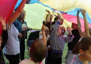 Grupa dzieci i dorosłych bawi się pod dużą, kolorową chustą animacyjną w barwach czerwonej, żółtej, niebieskiej i zielonej. Dzieci z uniesionymi rękami radośnie uczestniczą w zabawie, niektóre z nich mają pomalowane twarze. Wśród uczestników widoczne są również osoby dorosłe wspierające zabawę. Tło stanowi trawa i drzewa, a atmosfera jest radosna i integracyjna.