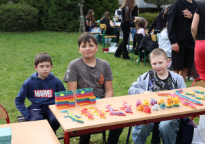 Na zdjęciu trzech chłopców siedzi za stołem ustawionym na trawie, prezentując kolorowe zabawki na szkolnym kiermaszu. Na stole znajdują się m.in. gumowe jaszczurki, figurki zwierząt oraz tęczowe pojemniki z ceną 10 zł. W tle widać inne osoby – dzieci i dorosłych – uczestniczących w wydarzeniu. Atmosfera jest spokojna i towarzyska, a całość odbywa się na świeżym powietrzu w otoczeniu zieleni.