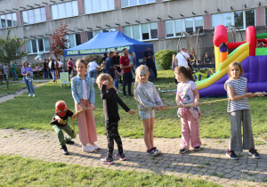 Na zdjęciu grupa dzieci przeciąga linę podczas pikniku szkolnego. Wśród uczestników jest chłopiec w masce Spider-Mana, dziewczynka z pomalowaną twarzą oraz inne dzieci ubrane kolorowo i uśmiechnięte. W tle widać dużą, kolorową dmuchaną zjeżdżalnię, niebieski namiot z napisem „Park Trampolin Air Jump” oraz osoby uczestniczące w wydarzeniu. Z tyłu znajduje się budynek szkolny. Atmosfera jest radosna i pełna dziecięcej energii.
